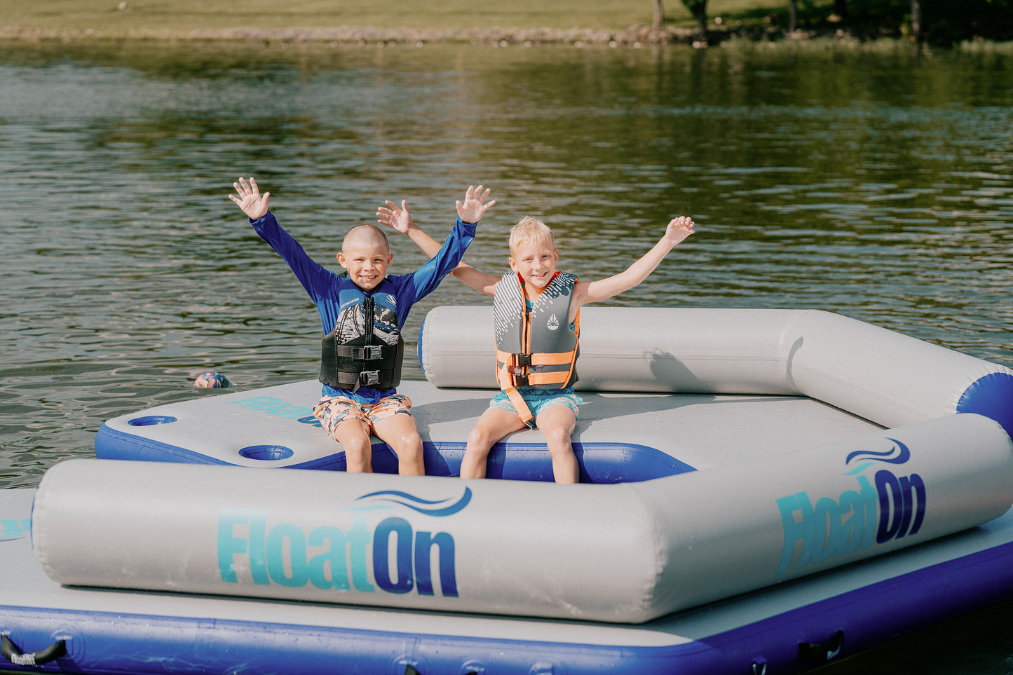 Two kids with their hands in the air sitting on a water mat from Float On.